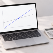 A laptop showing intersecting cost and revenue lines beside a calculator and notebook.